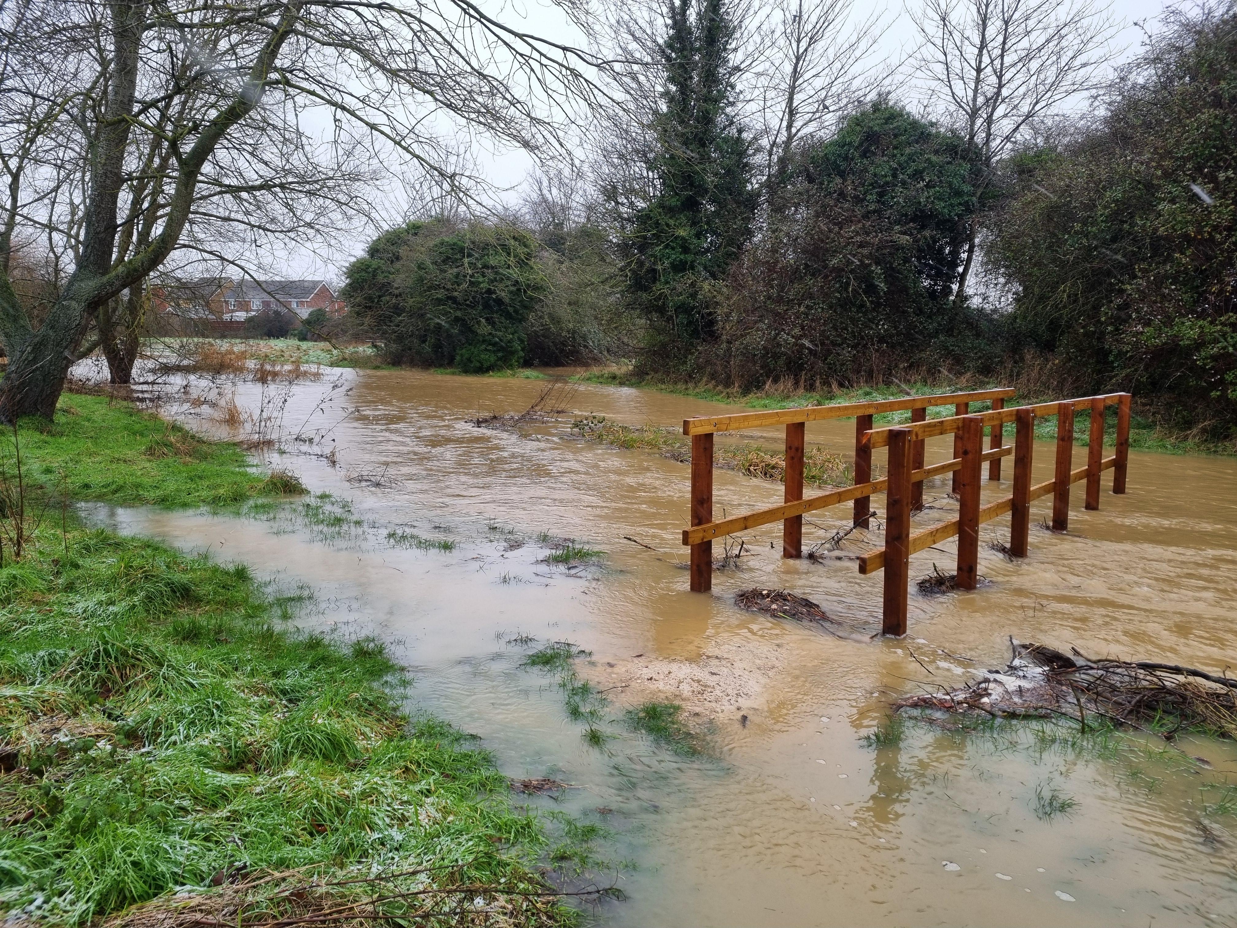 A bridge spans a flooded river, framed by a wooden fence, illustrating the impact of rising water levels.