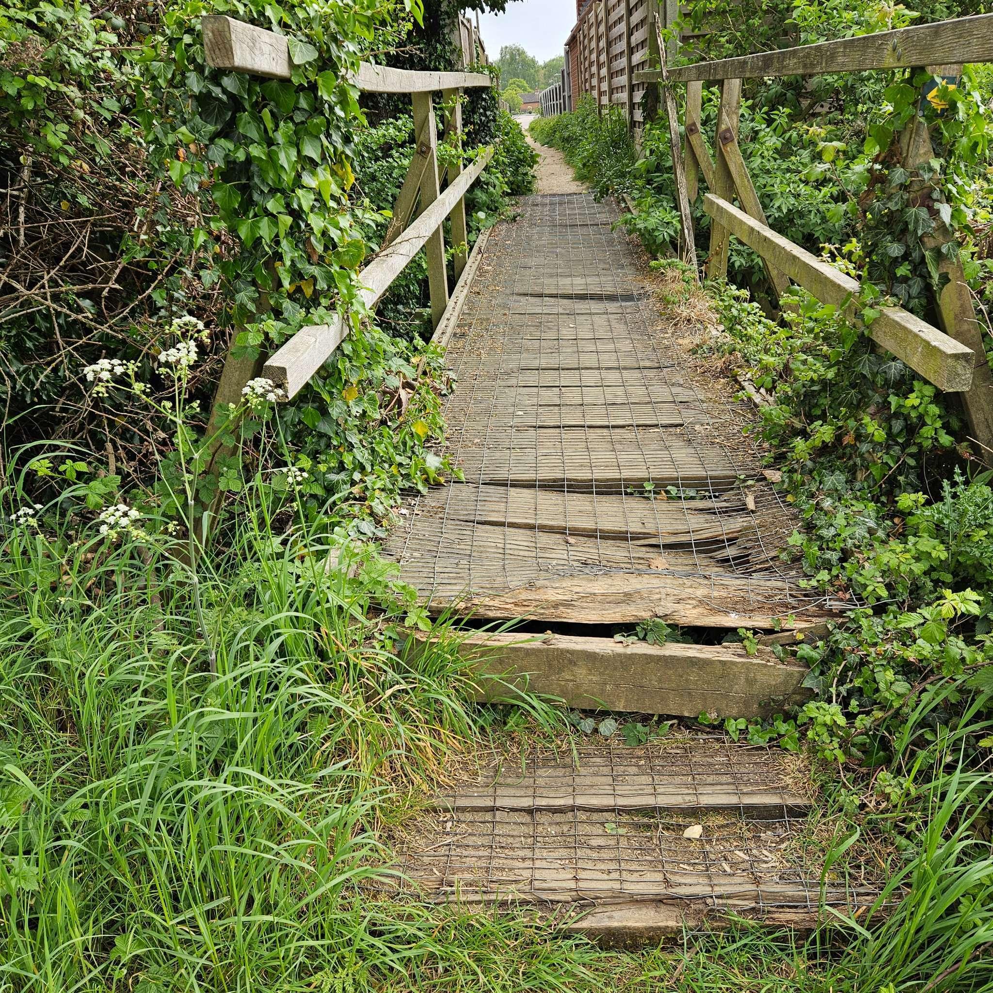 An unsafe wooden bridge spans a stream, surrounded by a lush green field under a clear blue sky.