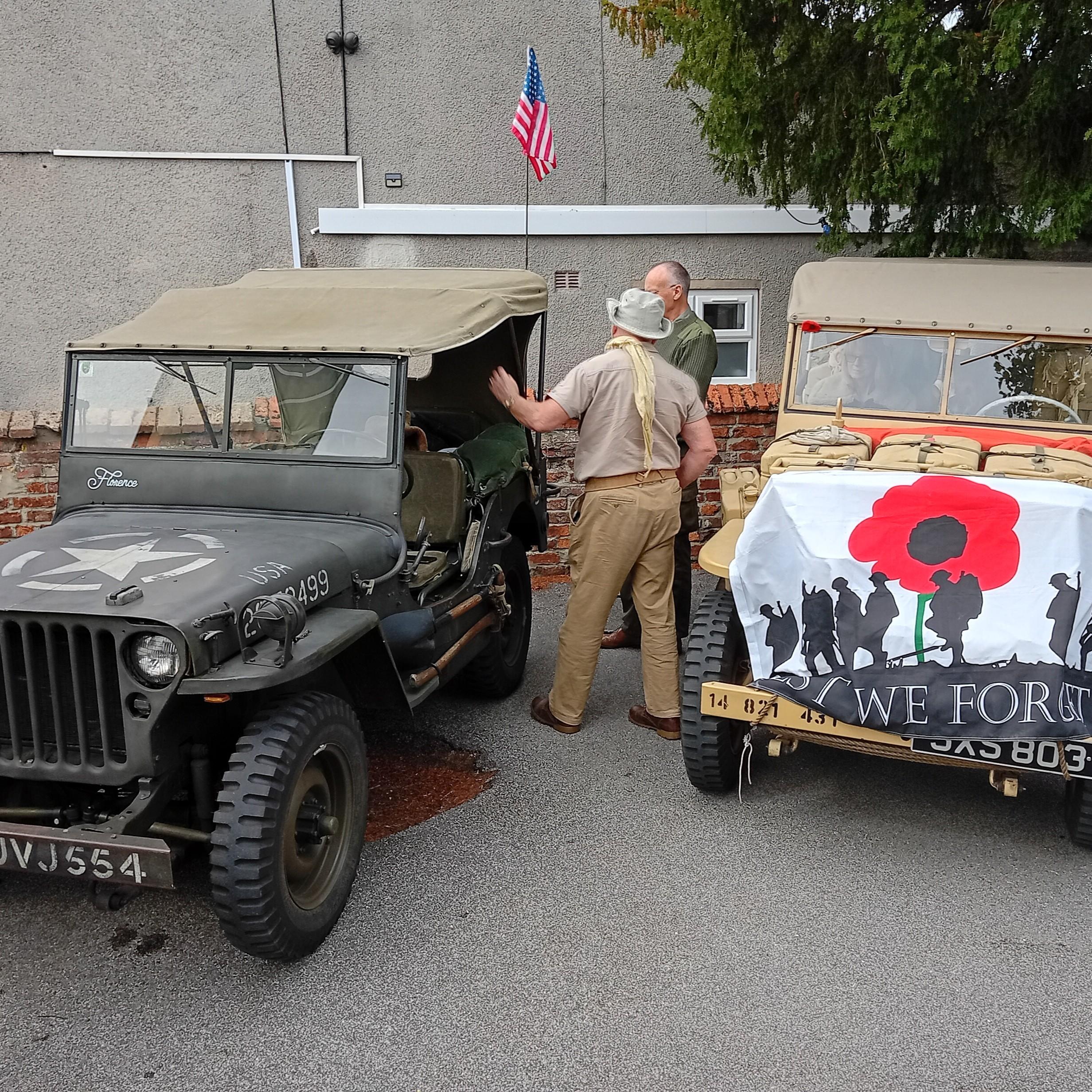 Two men stand beside a jeep adorned with a poppy, symbolizing remembrance and honoring fallen soldiers.