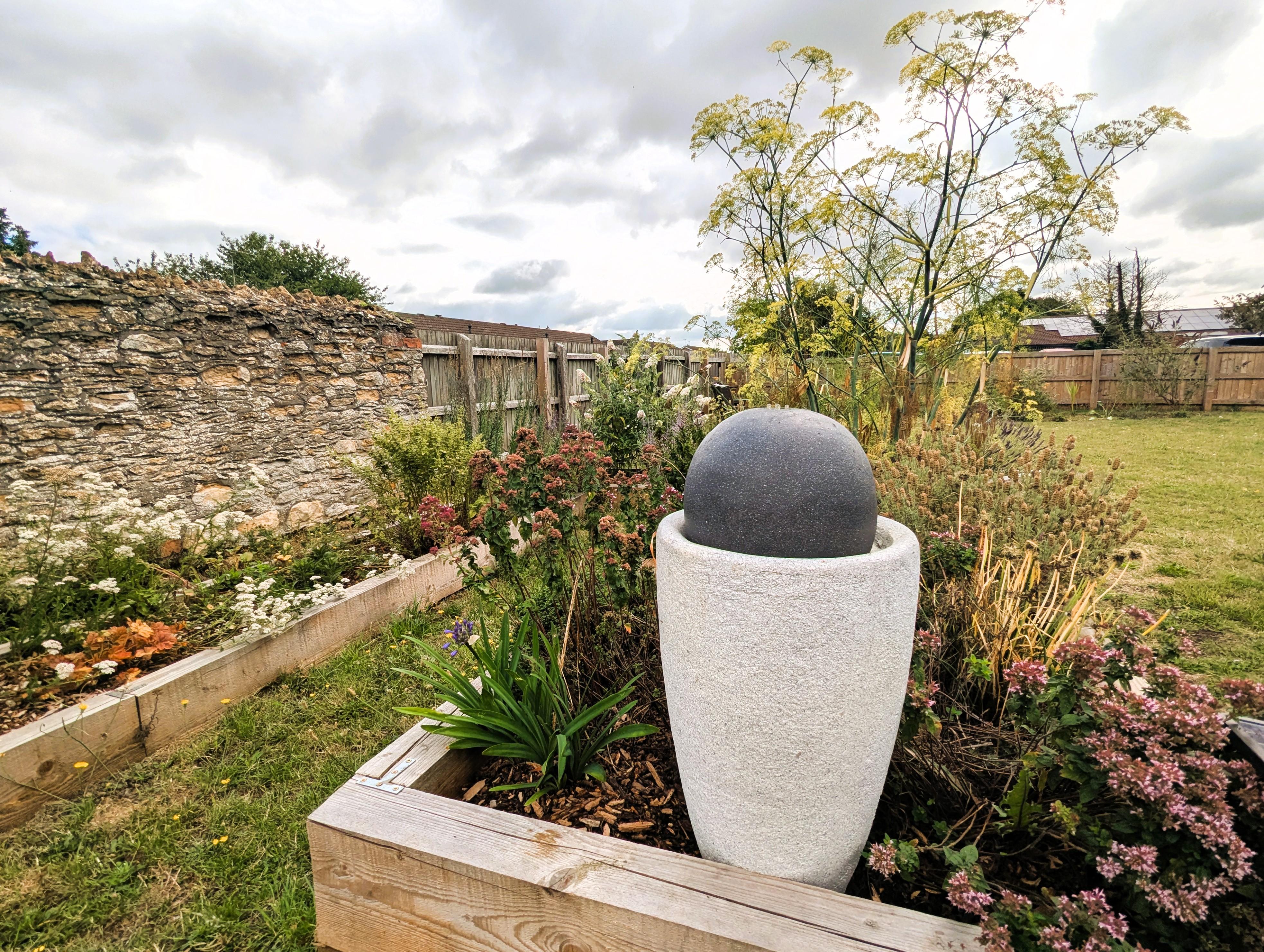 In a garden, a white vase stands against a stone wall, adding elegance to the lush greenery surrounding it.