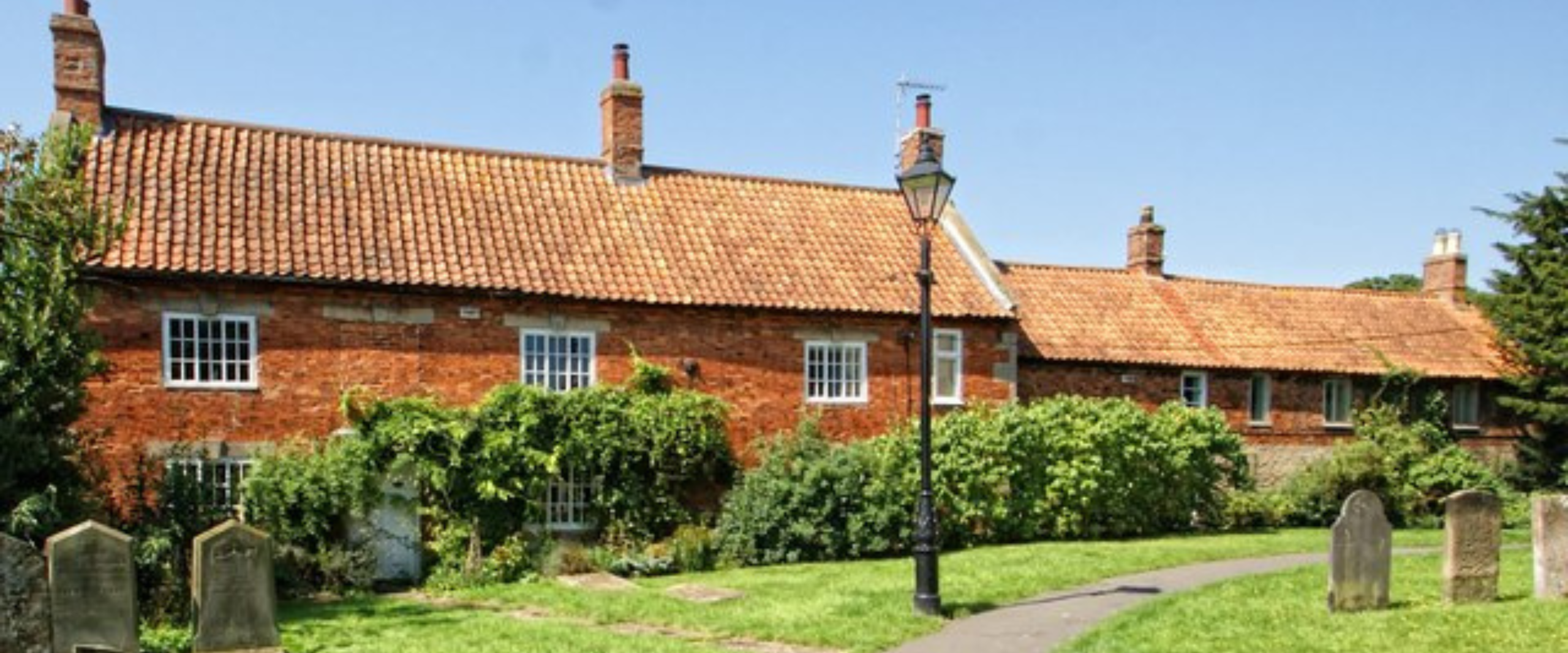 A red brick house with a white roof stands beside a neatly paved path, surrounded by greenery.