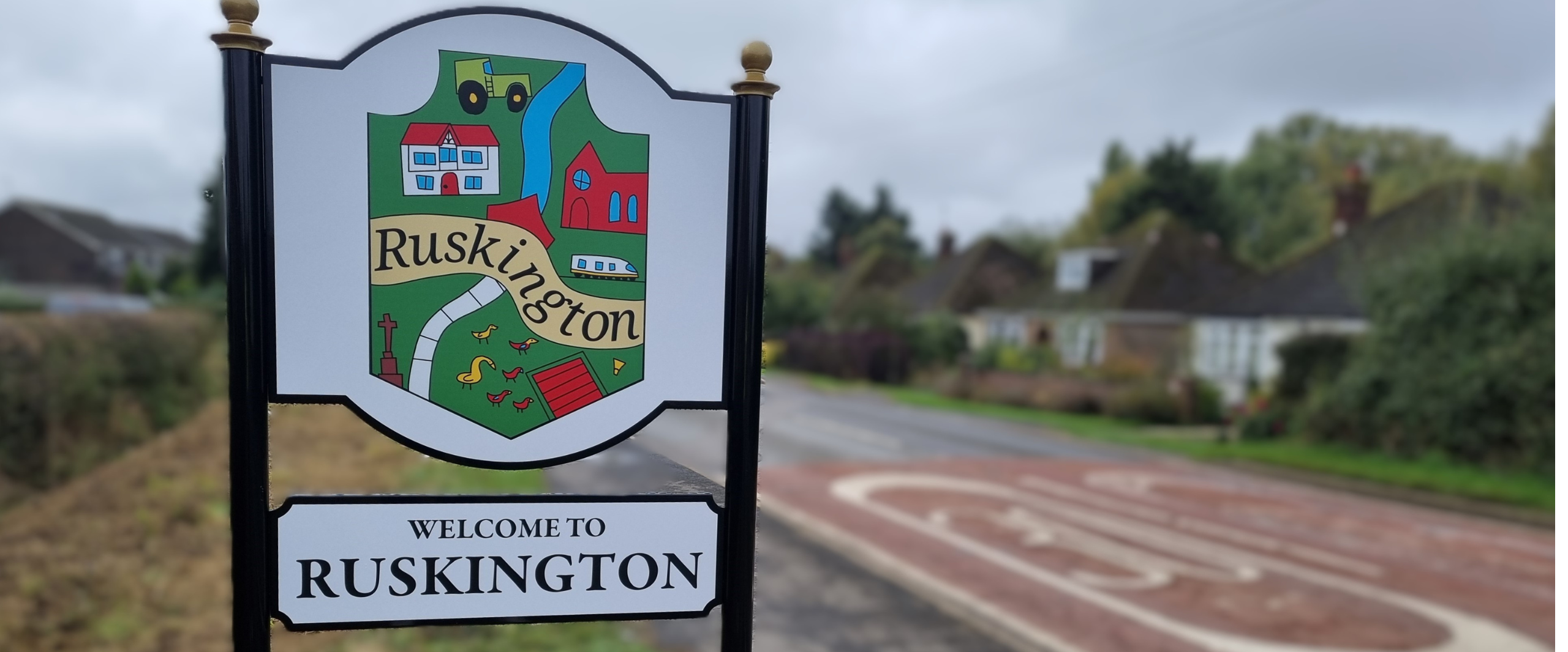 A charming welcome sign for Ruskington, displaying the village's name prominently with a picturesque landscape behind it.